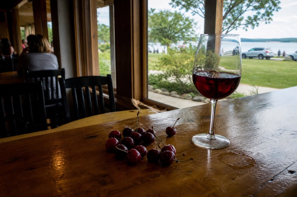 A glass of deep red fruit wine on a tasting room bar beside fresh cherries