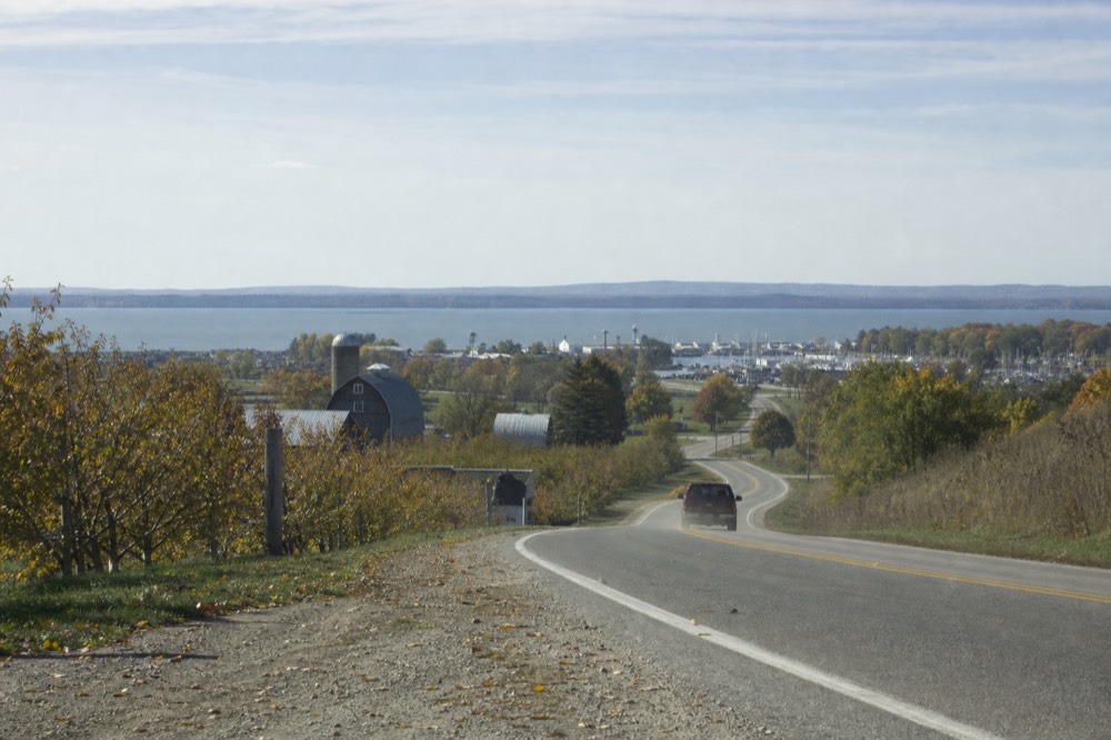 A two-lane lakeshore road curving past farmland toward a harbor village