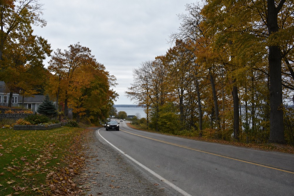 A two-lane road curving through dense autumn hardwoods with a sliver of lake beyond the trees
