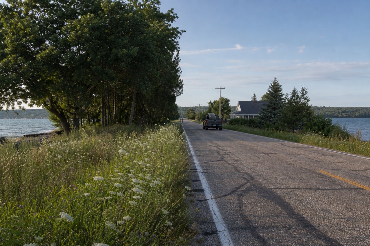 A narrow two-lane road running along a green peninsula with blue bay water on both sides