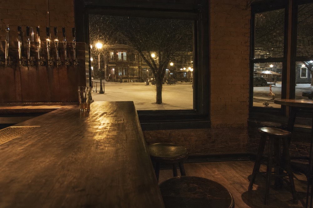 A brewery interior with warm lighting over a bar and empty stools at night