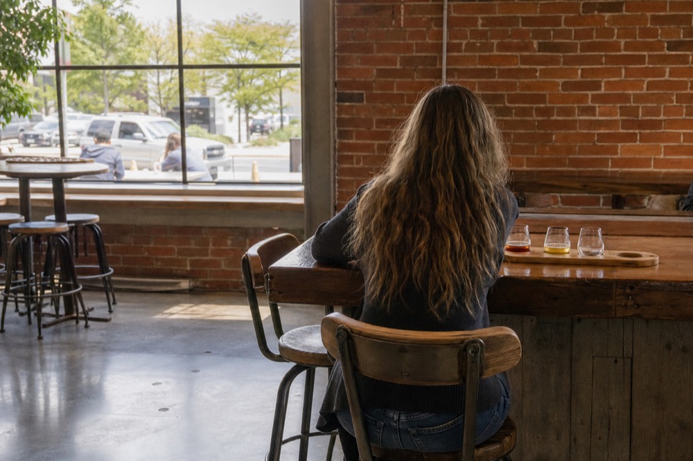 A single visitor standing at a wooden tasting bar with a flight of wine glasses