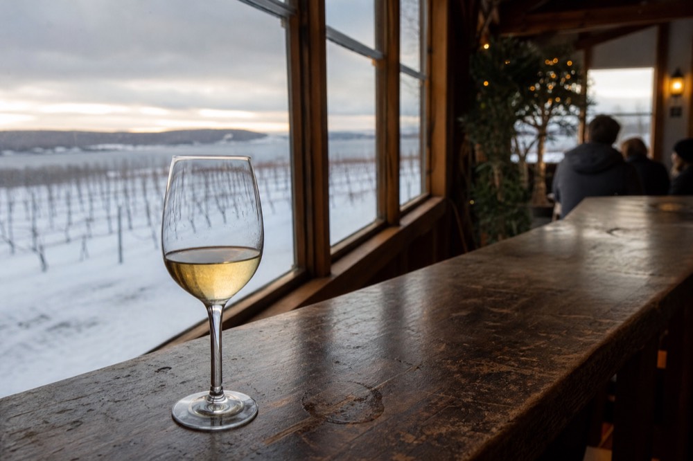 A glass of pale dessert wine on a tasting bar with snowy vineyards behind